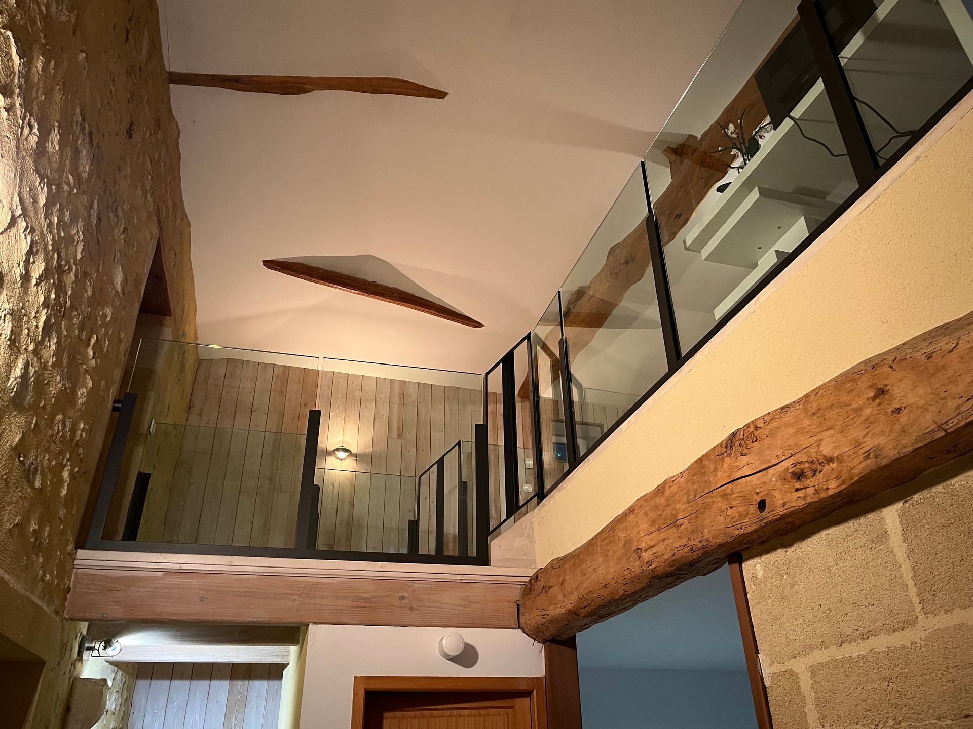 Intérieur moderne avec hauts plafonds, poutres apparentes, balustrade en verre et murs en pierre et plâtre.