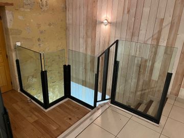 Une balustrade en verre à cadre métallique noir encadre un escalier, adossée à un mur en lattes de pierre et de bois, dans une pièce moderne.
