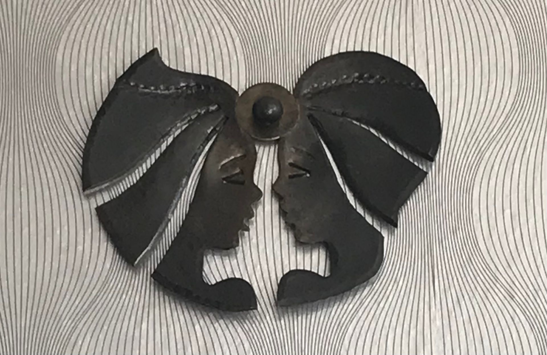 Une œuvre d'art murale symétrique en métal noir représentant deux profils se faisant face, reliés par un ornement circulaire central.