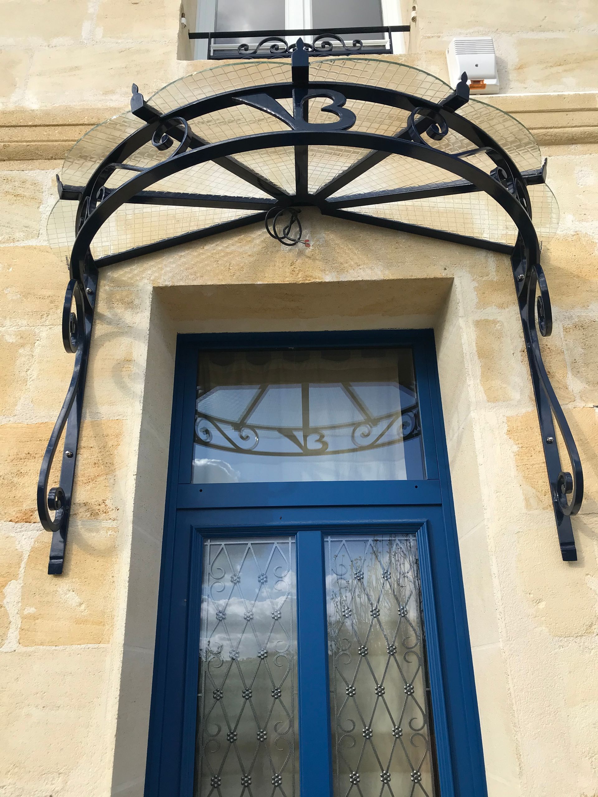 Une porte bleu foncé vitrée, surmontée d'un auvent arqué en métal noir orné, sur une façade en pierre claire.