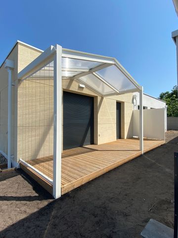 Une pergola à structure blanche et au toit translucide surplombe une terrasse en bois à motifs, adossée à un mur beige de la maison.
