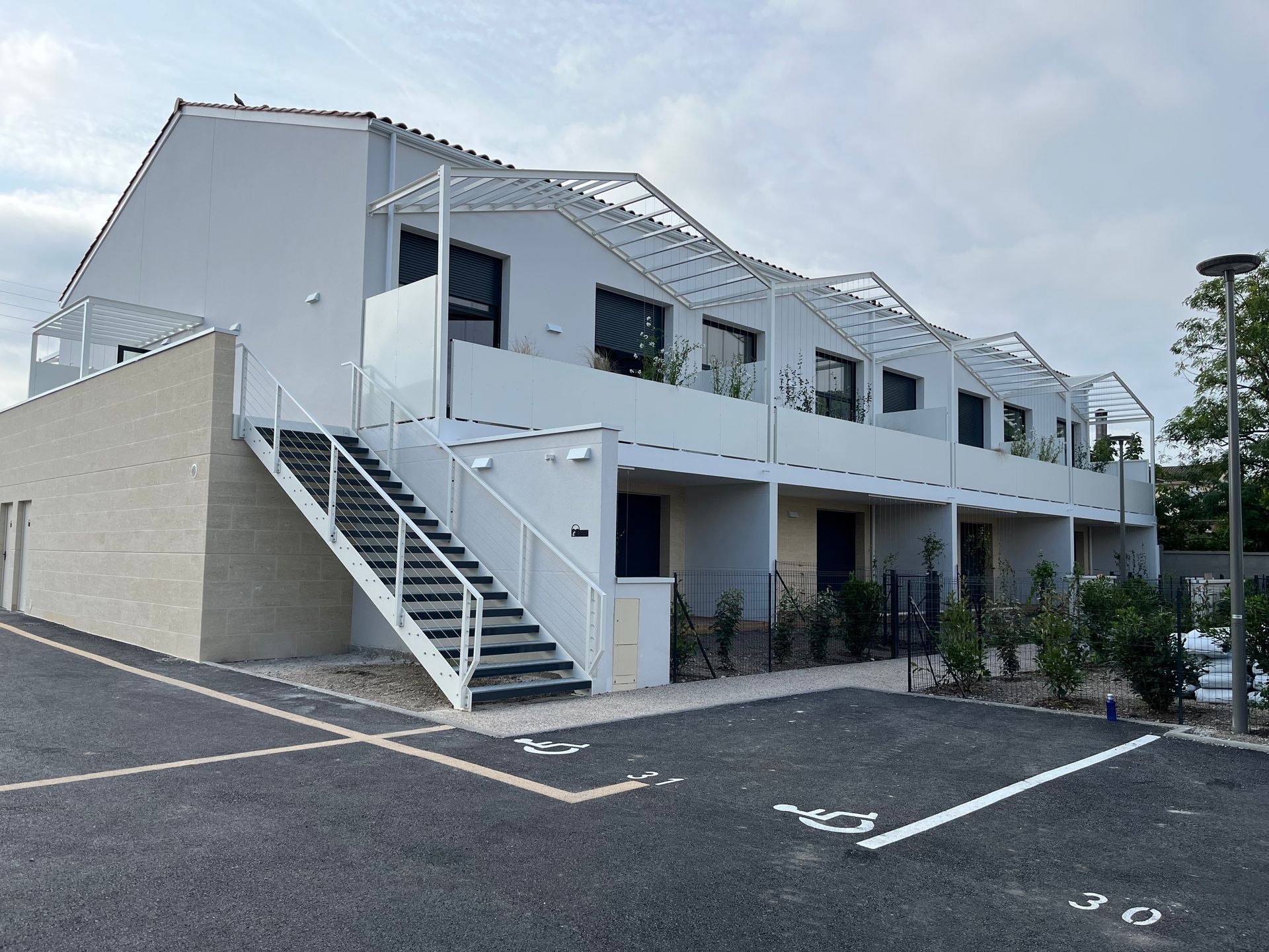 Immeuble résidentiel blanc à plusieurs logements, avec un escalier extérieur en métal et des places de stationnement réservées aux personnes à mobilité réduite.