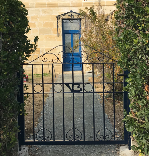Une clôture et un portail en métal gris foncé à claire-voie, fixés sur un mur en béton blanc devant une maison.