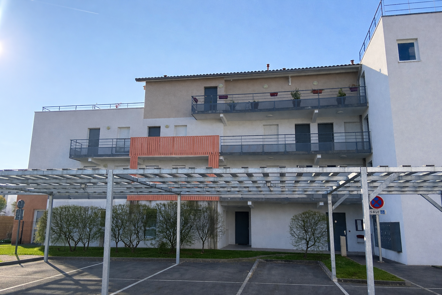 Un immeuble d'appartements blanc à plusieurs étages avec des balcons et un abri de parking métallique au premier plan.