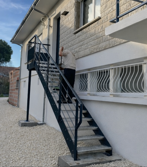 Une personne monte un escalier en métal noir à côté d'une maison en pierre.