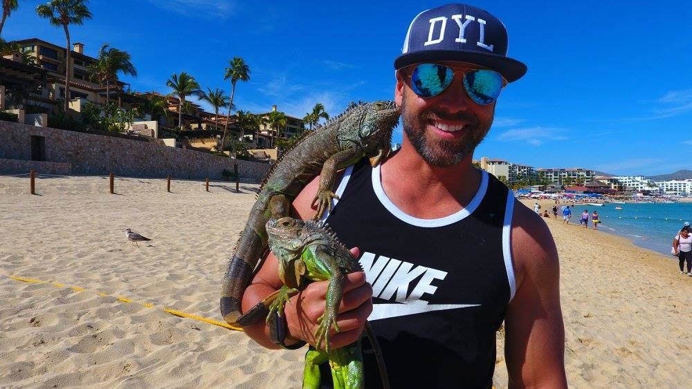 Man standing on steps, smiling, holding food, facing a table of people at a pool-side dinner party.