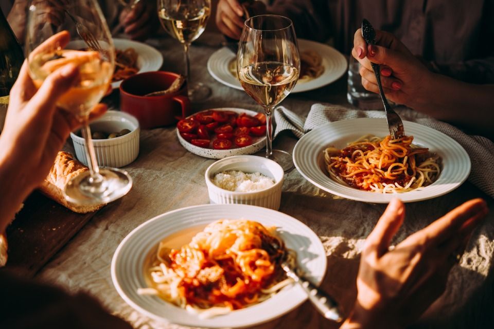People dining at a table with pasta, wine, and side dishes. Hands reach for food and drinks.
