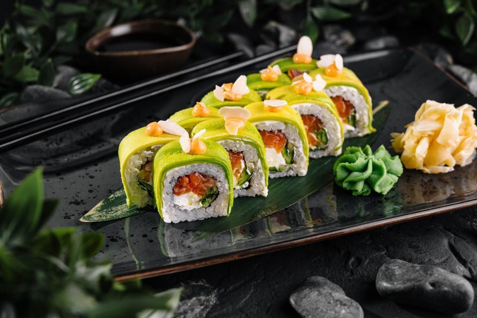 Sushi rolls with avocado and shrimp, on a black plate, with chopsticks and condiments.