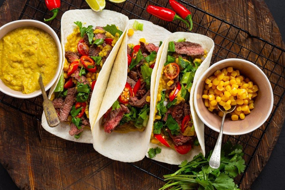Steak tacos with toppings and sides of sauce and corn on a wooden board.