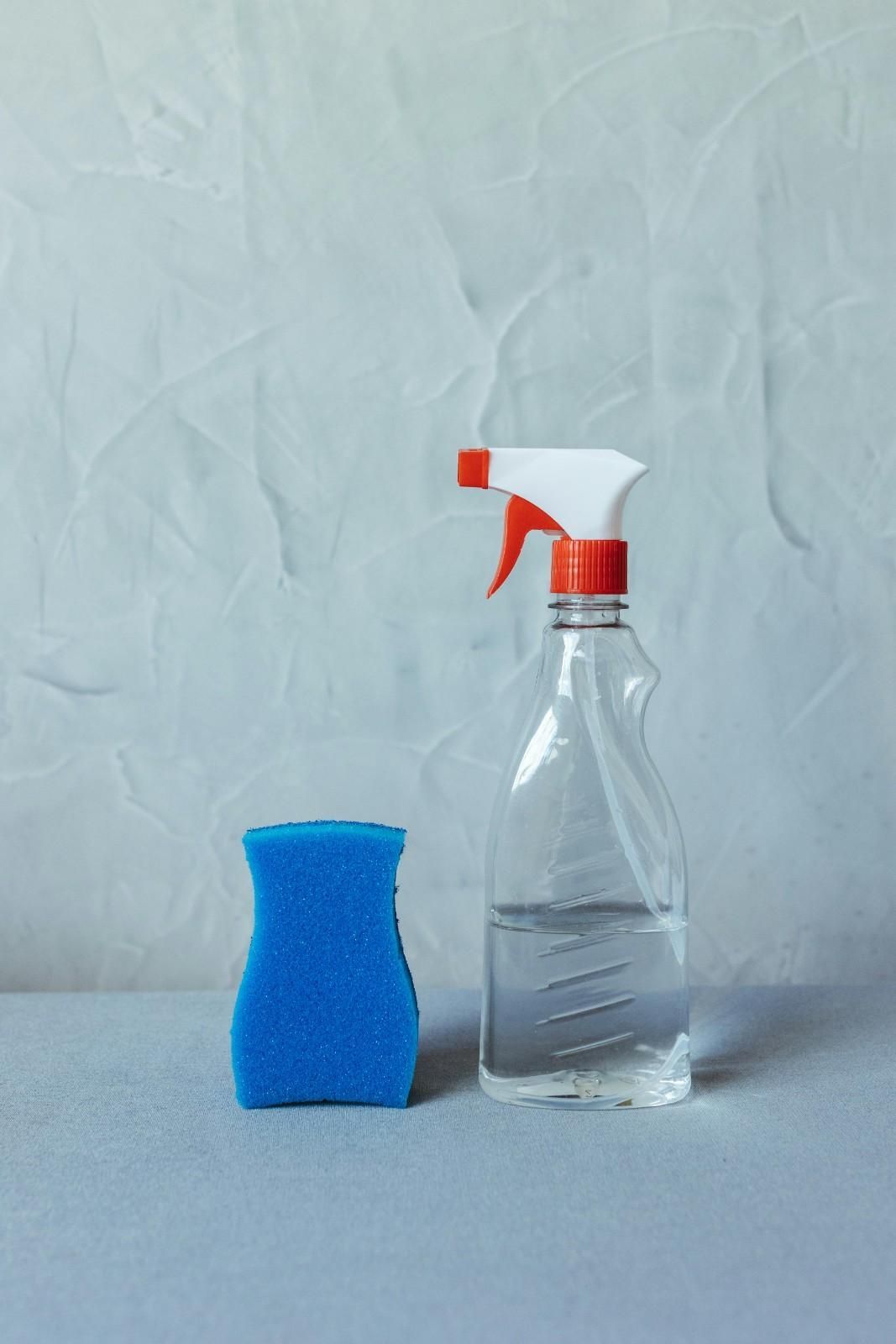 Esponja azul y botella de spray con solución limpiadora sobre una superficie gris sobre un fondo texturizado.