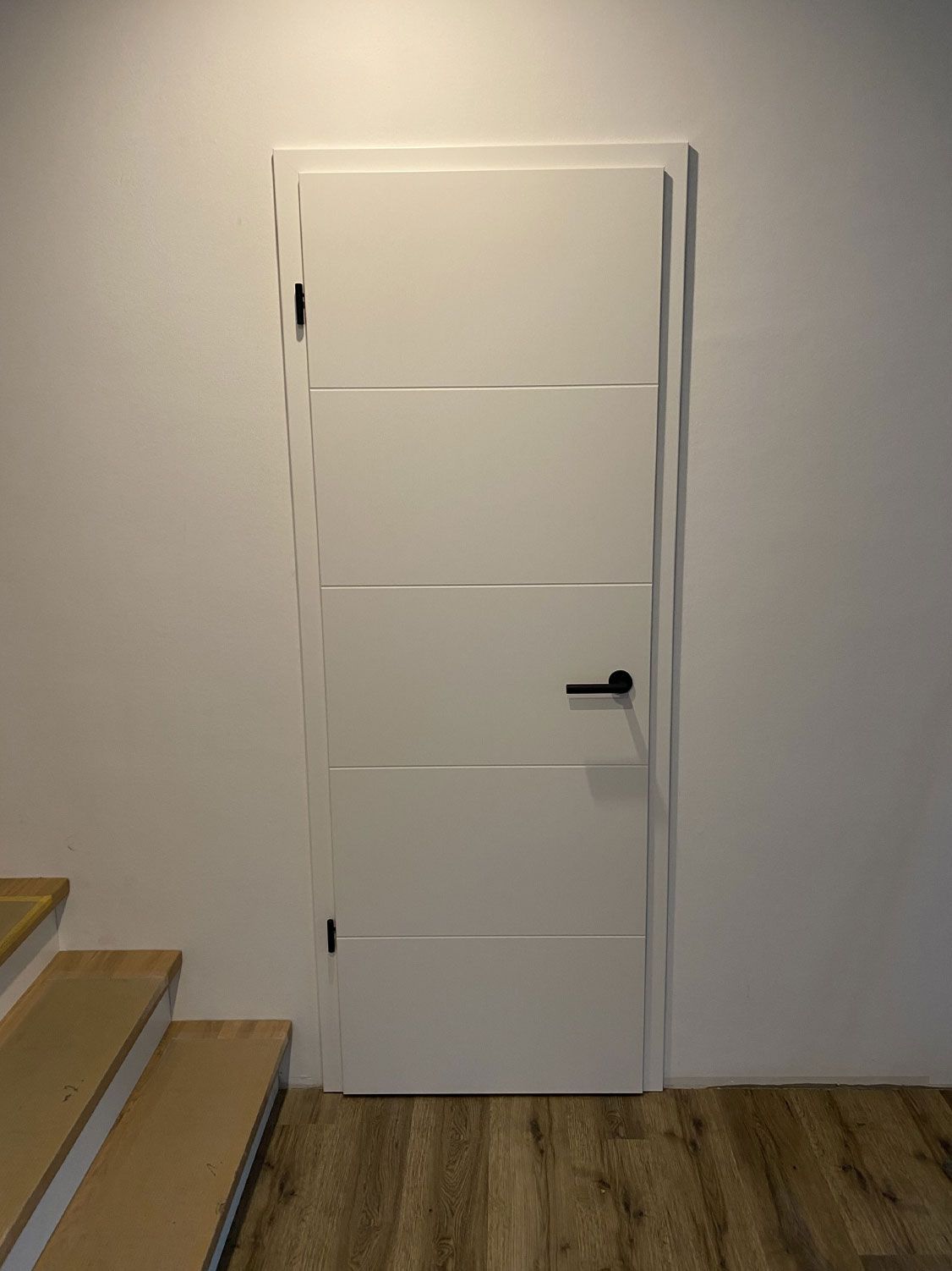In einem Flur neben einer Treppe befindet sich eine weiße Tür mit einem schwarzen Griff.