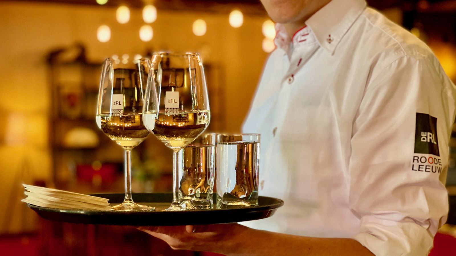 Kellner hält ein Tablett mit Weingläsern und Schnapsgläsern in einem warm beleuchteten Restaurant.
