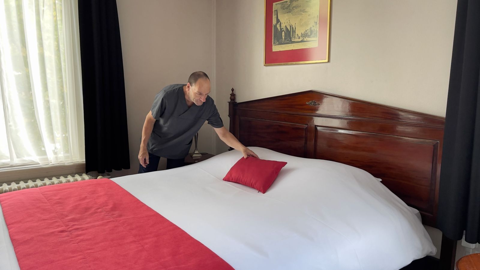Mann in einem Hotelzimmer rückt ein rotes Kissen auf dem Bett zurecht. Rote Decke und Kopfteil; Fenster mit Vorhang.