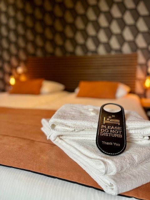 Bett mit orangefarbener Tagesdecke, weißen Handtüchern und „Bitte nicht stören“-Schild. Dekorative geometrische Tapete.