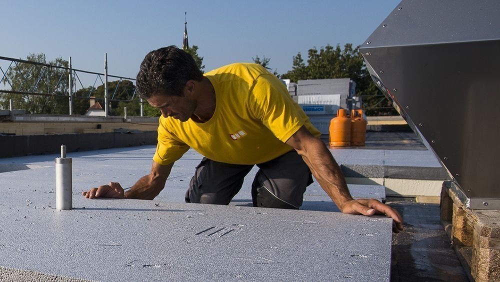 Un ouvrier en chemise jaune, agenouillé sur un toit plat, inspecte la surface blanche d'un toit.