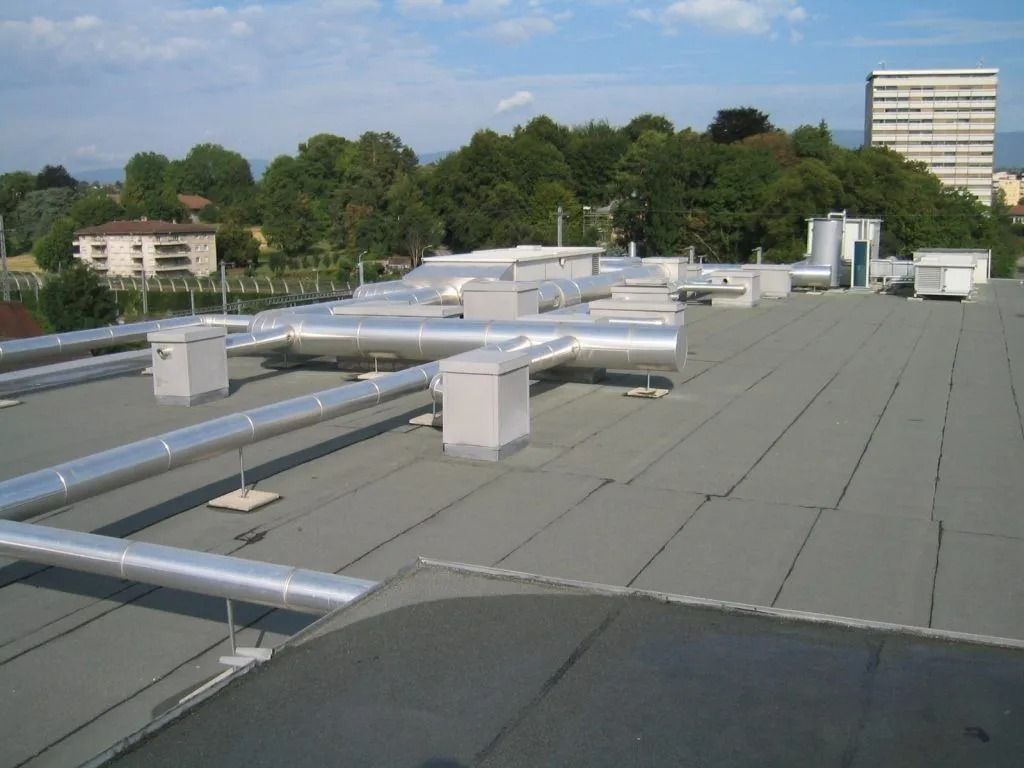 Toit avec conduits et aérations métalliques, surplombant une ligne d'horizon arborée sous un ciel bleu.