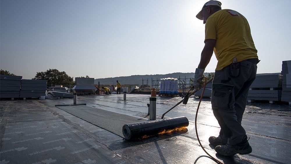 Un ouvrier utilise un rouleau pour lisser le matériau de couverture sur un toit plat au coucher du soleil.