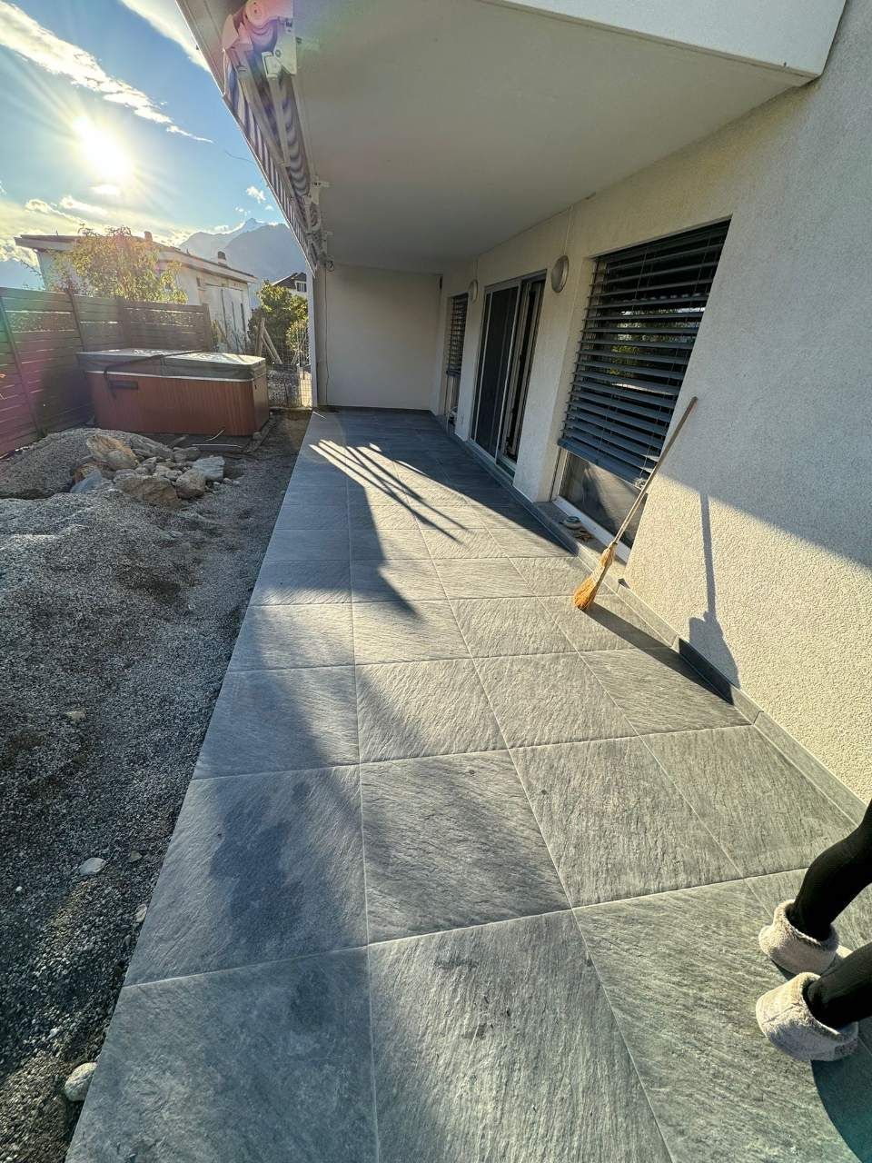 Terrasse carrelée de gris foncé, sous un auvent blanc. La lumière du soleil projette des ombres. 