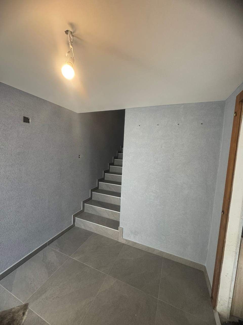 Vue intérieure : un escalier gris monte. Murs gris, sol carrelé clair et un unique plafonnier.