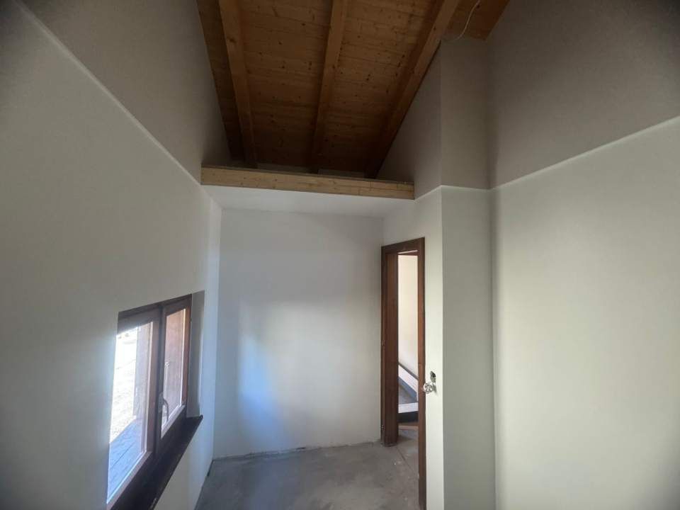 Couloir intérieur avec poutre en bois, fenêtre et porte ; les murs sont peints en blanc et gris.