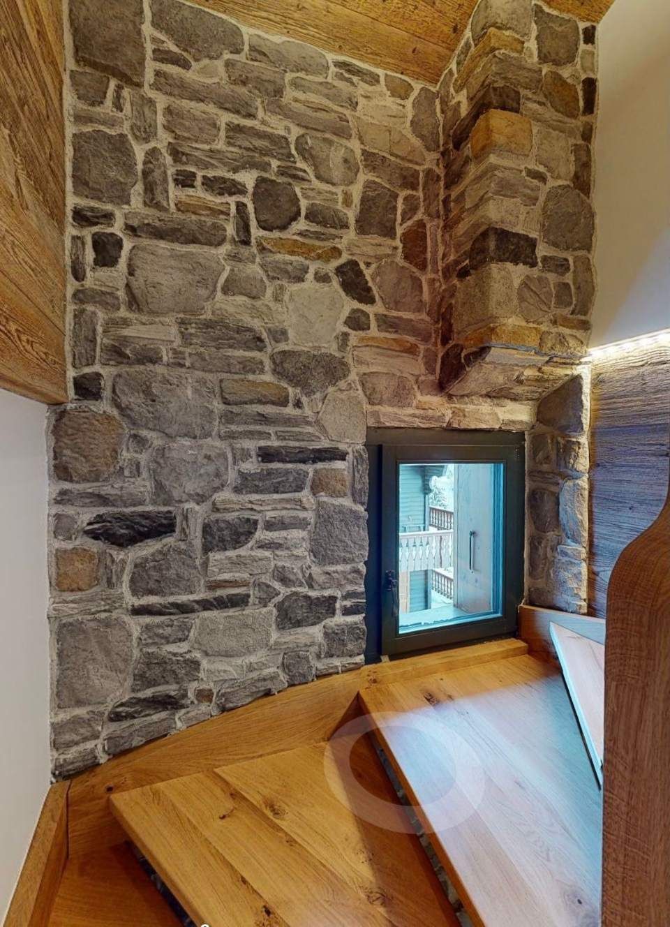 Mur en pierre avec une petite fenêtre près d'un escalier en bois, éclairé par un éclairage d'ambiance.