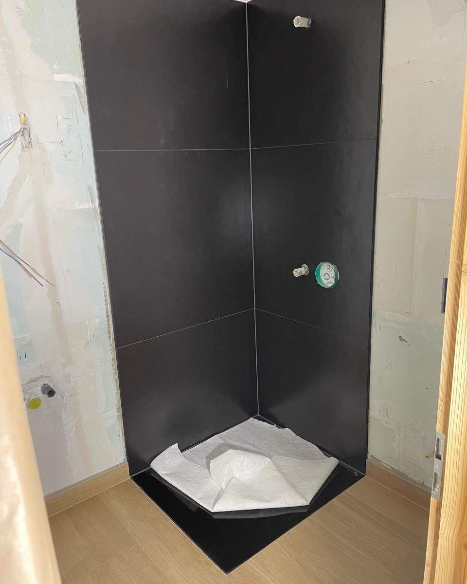 Cabine de douche d'angle carrelée de noir, avec murs non finis et protection de sol blanche.