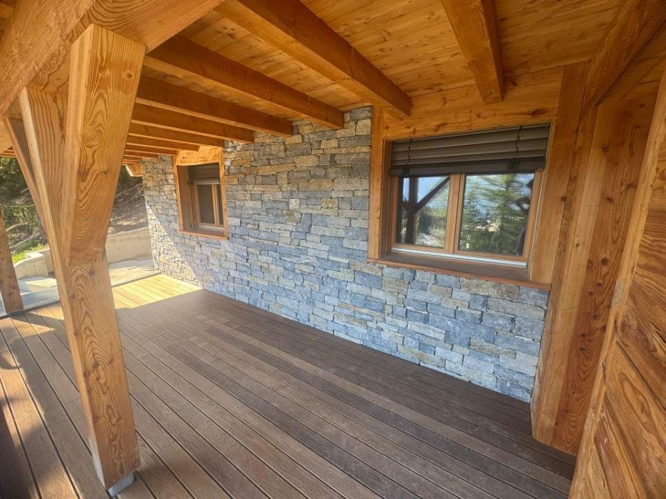 Véranda en bois avec mur en pierre, fenêtres et volets foncés ; terrasse et poutres brunes.
