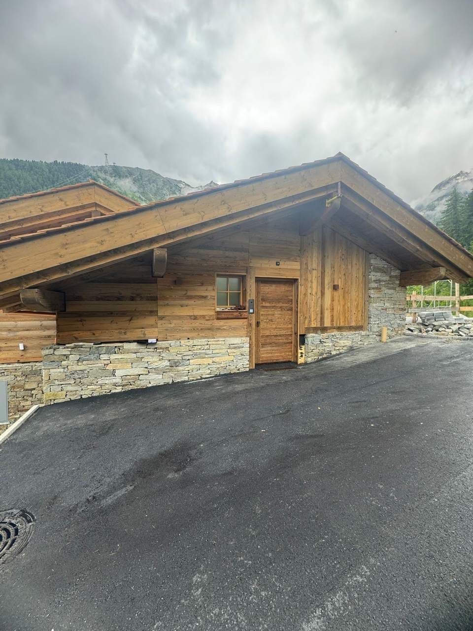 Chalet en bois avec soubassement en pierre, allée asphaltée et panorama montagneux sous un ciel nuageux.