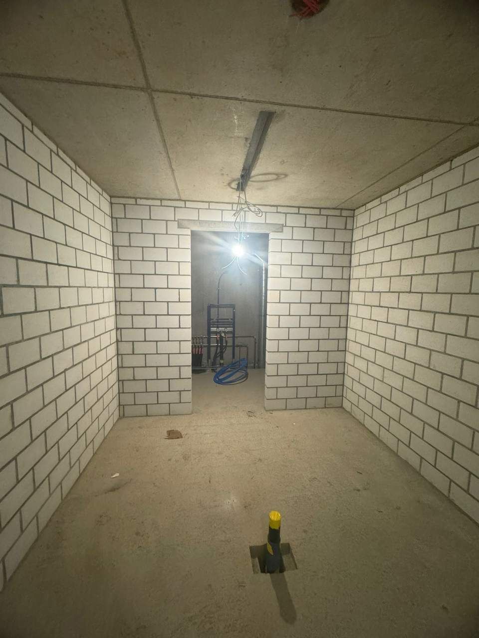 Pièce inachevée aux murs de briques grises, au sol en béton et avec une porte donnant sur une autre pièce. 