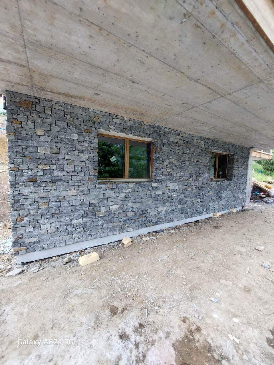 Mur extérieur en pierre avec deux fenêtres sous un plafond en béton. Chantier avec sol en terre battue.