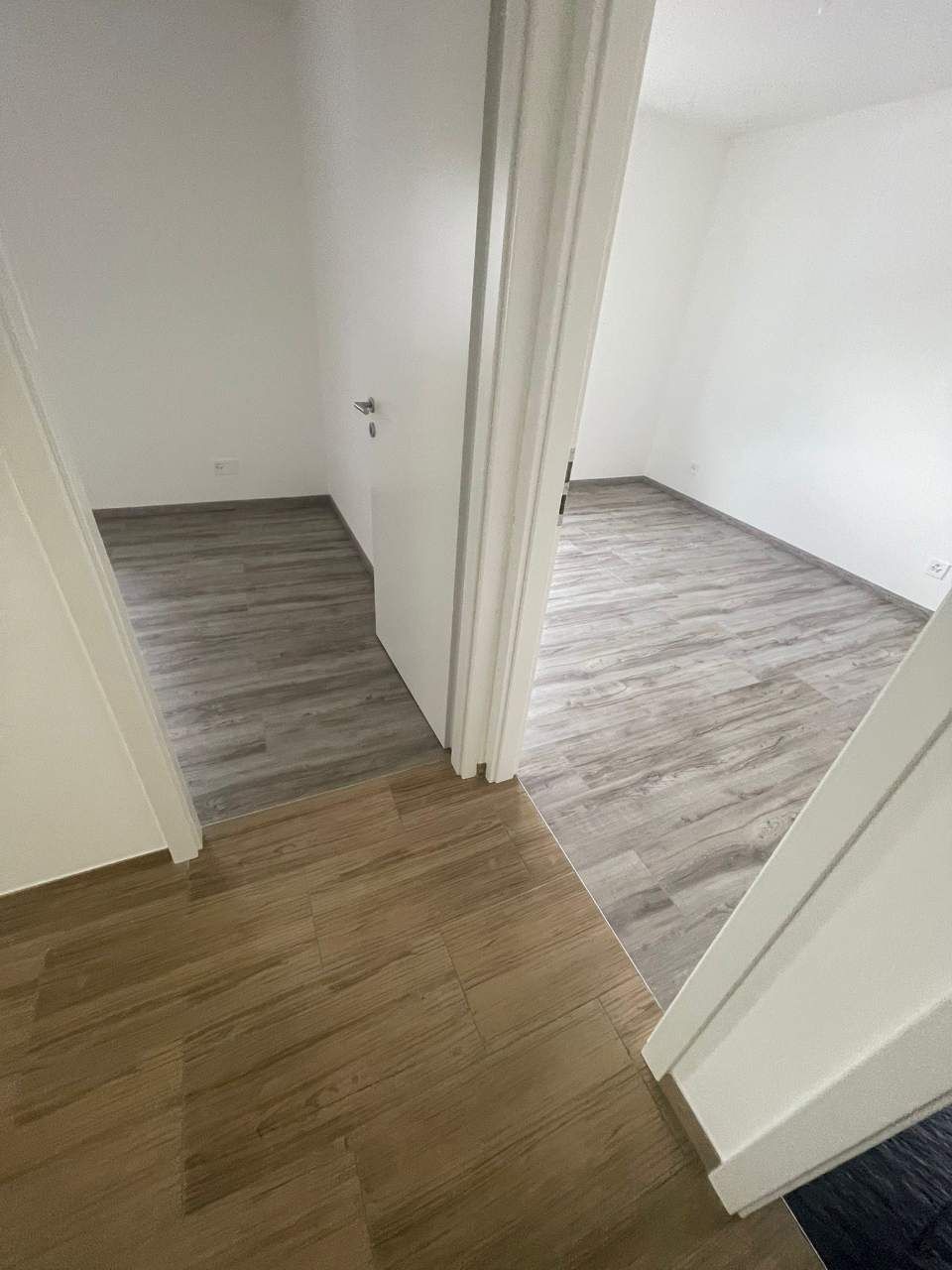 Vue depuis un couloir sur deux pièces avec un sol gris clair et imitation bois, des murs blancs et des portes ouvertes.