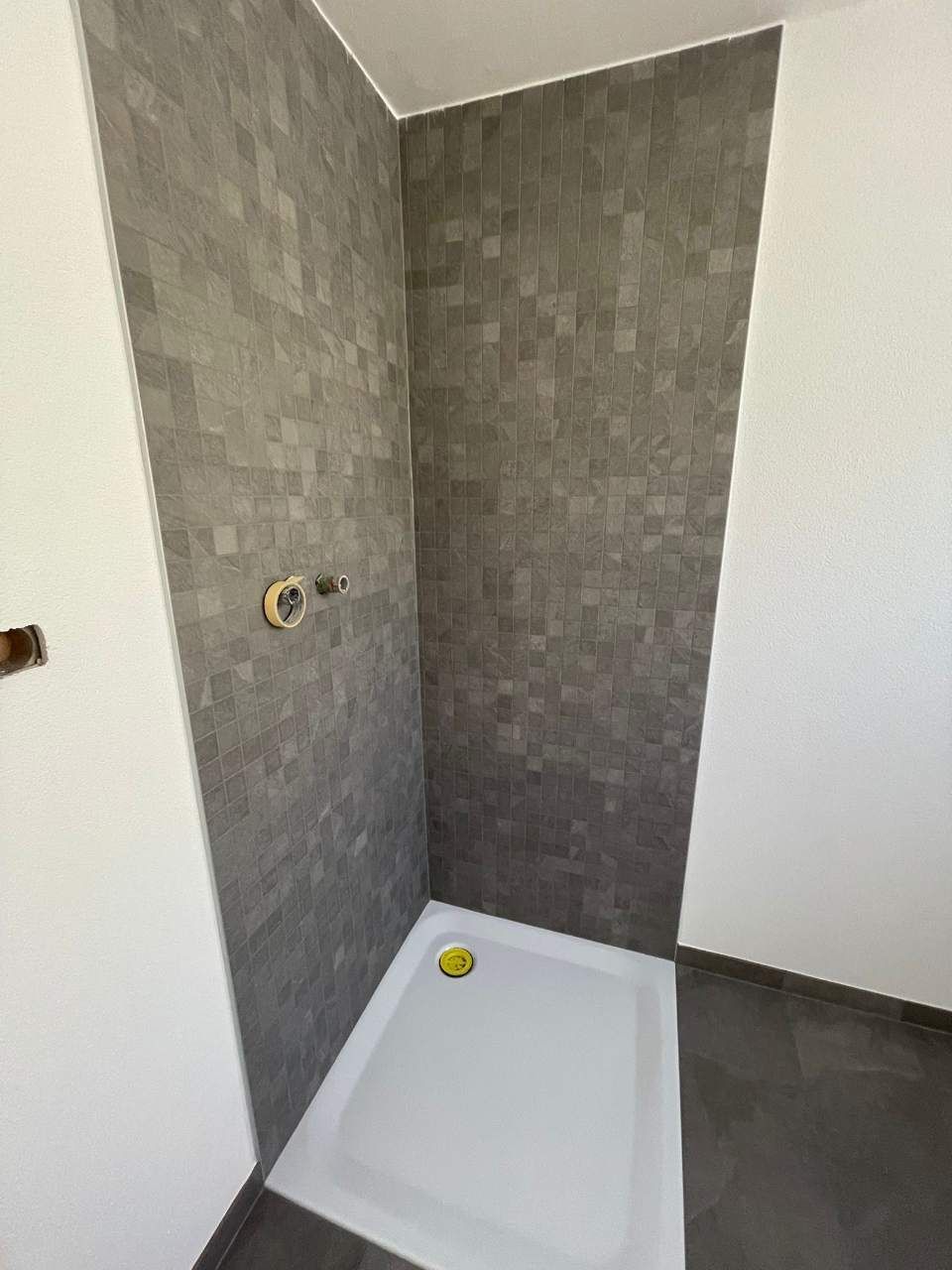 Cabine de douche avec murs carrelés gris, receveur de douche blanc et robinetterie.