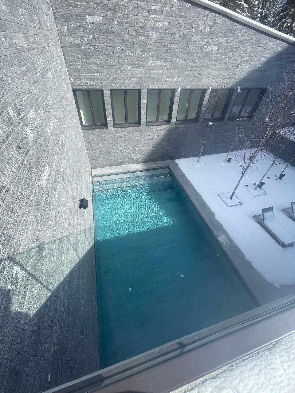 Vue extérieure d'une piscine rectangulaire entourée d'une terrasse enneigée. Murs et fenêtres en pierre grise.