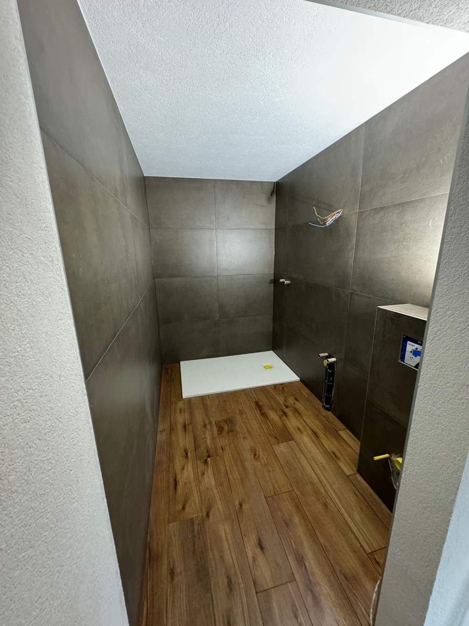 Douche aux murs carrelés de brun, au sol imitation bois et au plafond blanc. Un tapis de bain blanc est posé au sol.