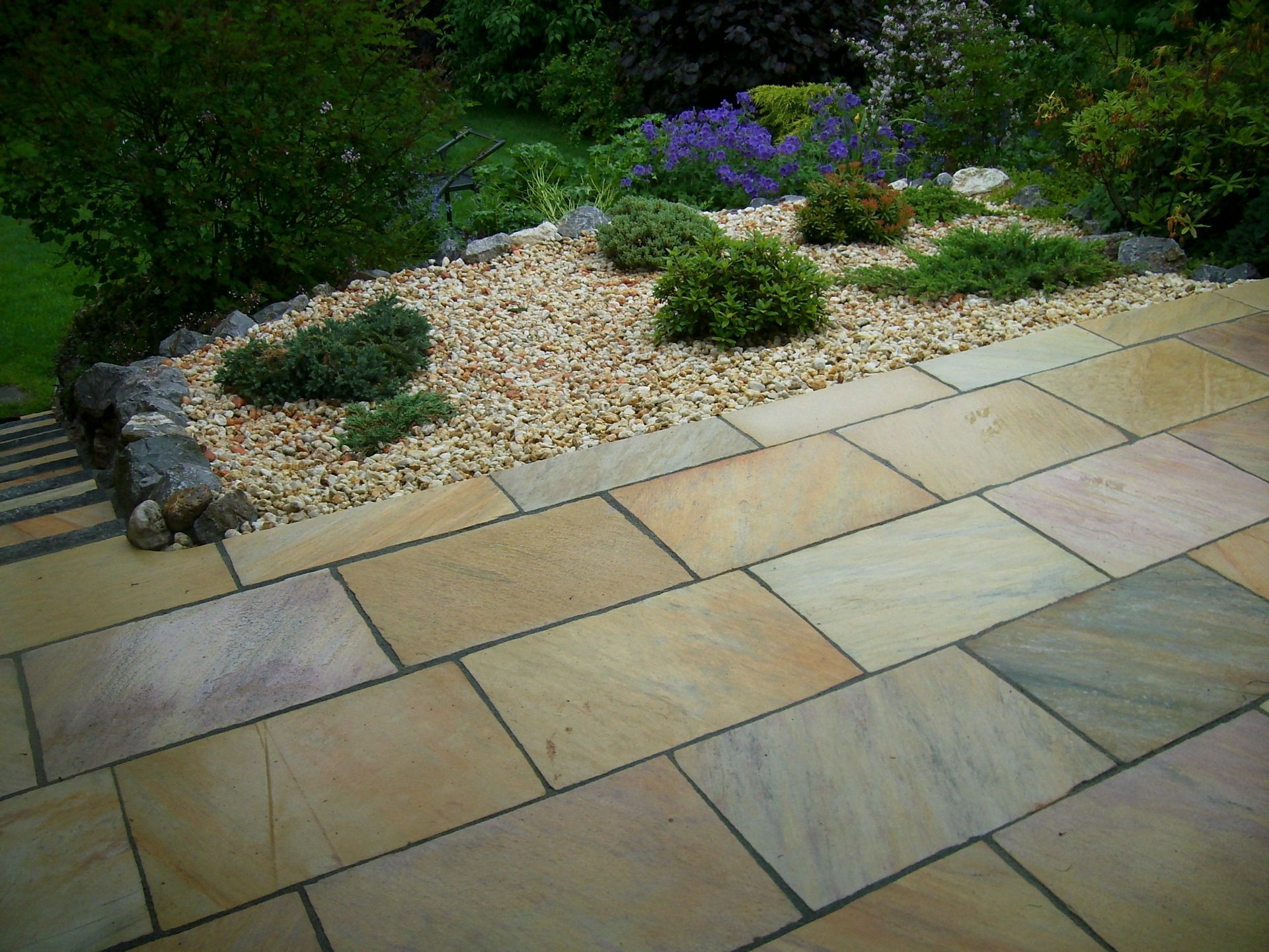 Terrassenbereich mit beigen Natursteinplatten, angrenzendem Kiesbeet und üppiger Gartenbepflanzung