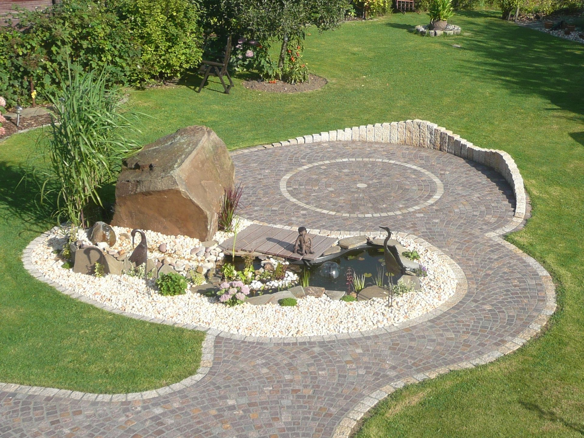 Gartenlandschaft mit einem runden Gartenteich, umgeben von Natursteinpflaster und einem großen Findling