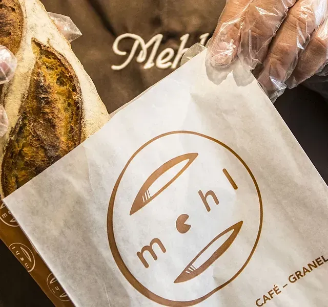 Una persona con guantes sostiene una barra de pan, parcialmente dentro de una bolsa de papel con el logotipo de una panadería.