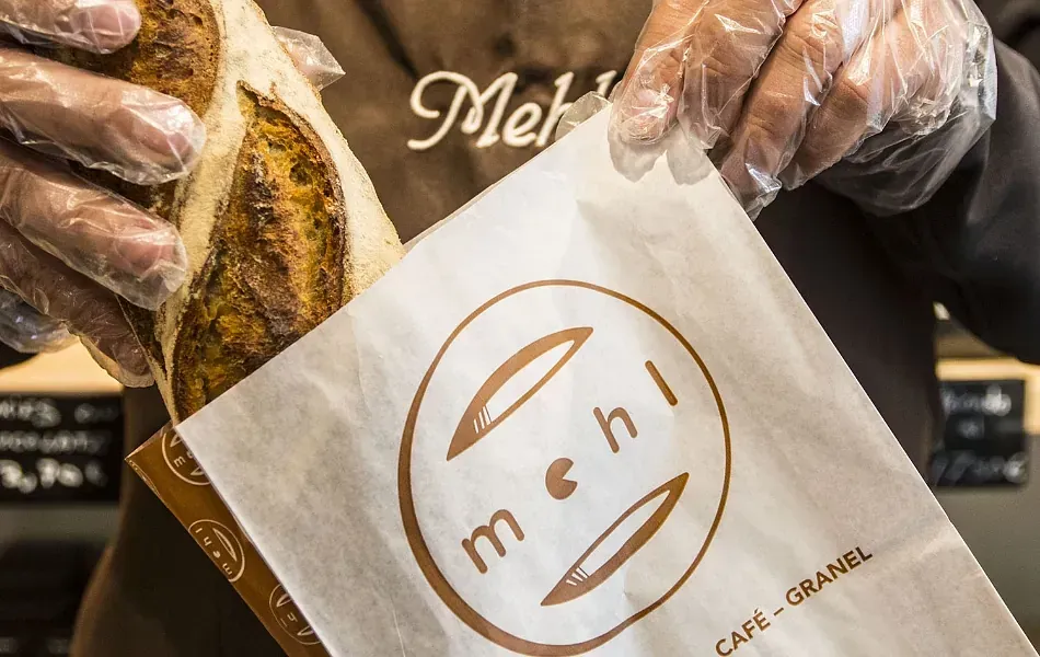 Persona con guantes colocando una barra de pan en una bolsa de papel con el logotipo de la panadería 