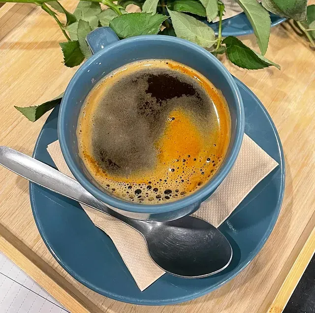 Una taza de café negro con crema reposa en una taza azul sobre un platillo azul, con una cuchara y una servilleta sobre una superficie de madera.