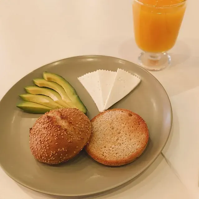 Plato con rodajas de aguacate, queso y bagel, servido con un vaso de jugo de naranja.