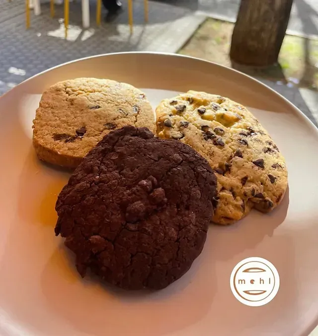 Taza de café con espuma y canela, un panecillo en un plato y gafas de sol sobre una mesa blanca.