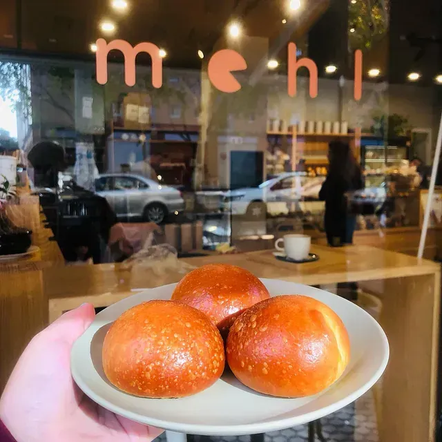 Una persona sostiene un plato con tres bollos dorados frente a la ventana de una panadería que muestra la palabra 