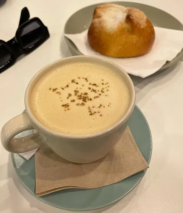 Taza de café con espuma y canela, un panecillo en un plato y gafas de sol sobre una mesa blanca.