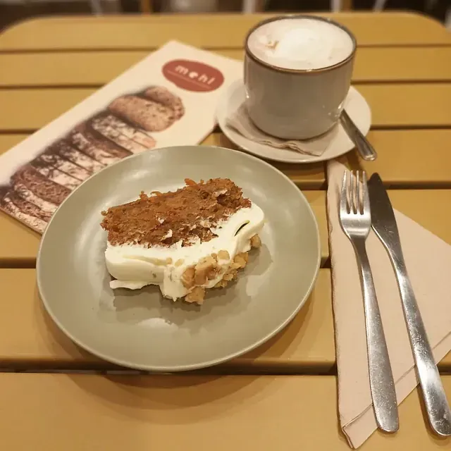 Una rebanada de pastel de zanahoria en un plato, café y un menú sobre una mesa. También hay cubiertos y una servilleta.