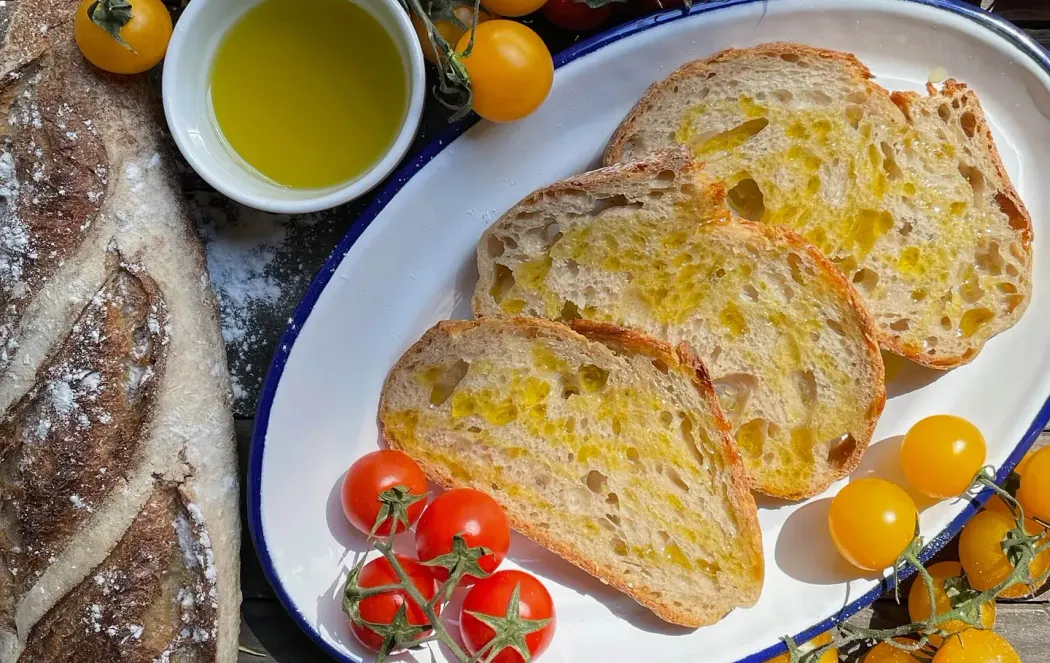 Rebanadas de pan rociadas con aceite de oliva, servidas con tomates cherry y un tazón de aceite de oliva en un plato blanco.