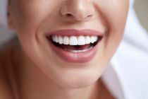 Close da boca de uma pessoa, língua tocando os dentes com aparelho. Piercing no nariz visível; sorrindo.