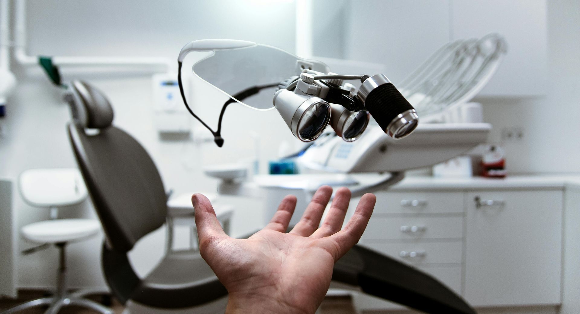 Uma mão estendida em direção a uma cadeira odontológica e lupas cirúrgicas em um consultório limpo e iluminado.