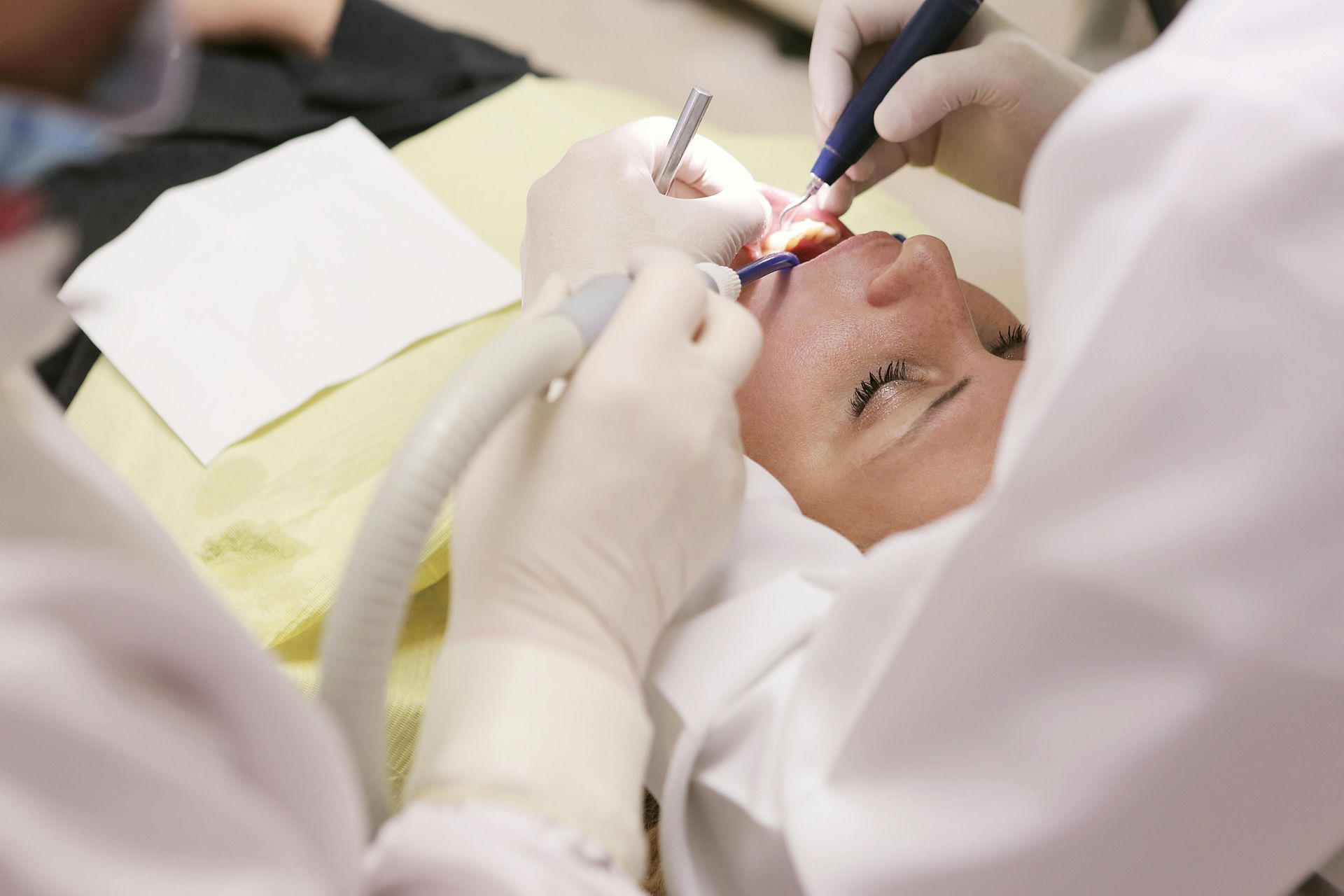 Mulher recebendo tratamento odontológico de dentista e assistente vestindo jalecos brancos.