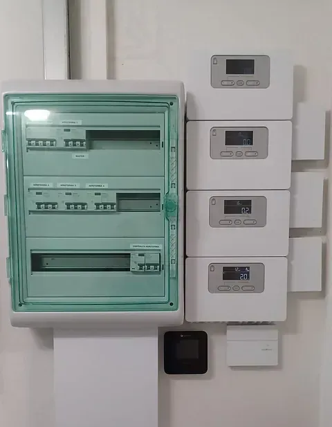Una caja eléctrica verde está sobre una pared blanca al lado de una fila de cajas blancas.