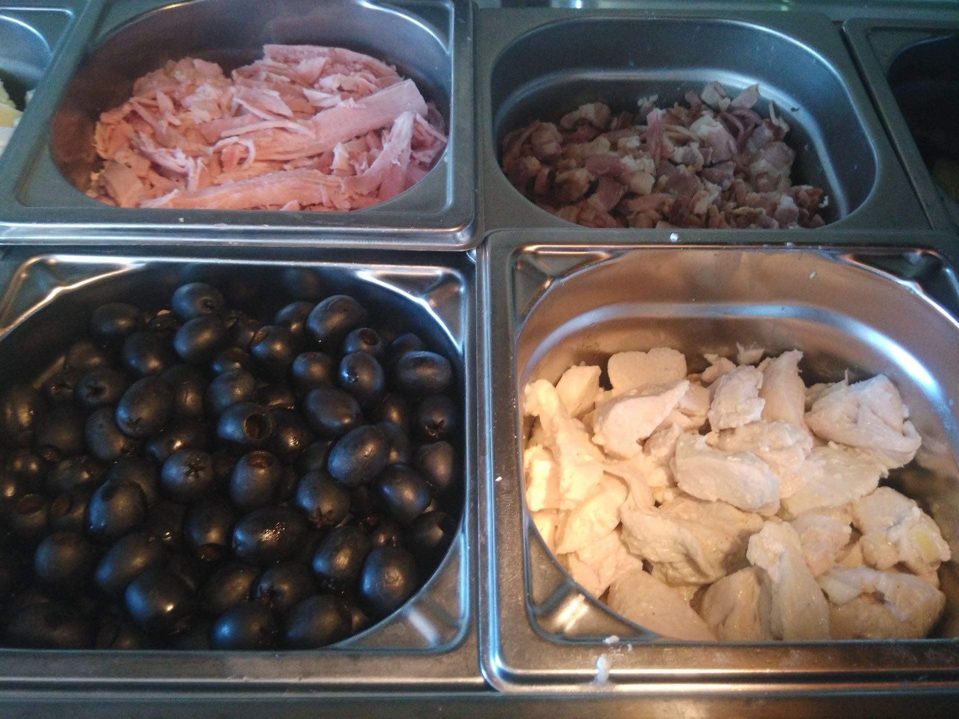 Quatre plateaux de nourriture contenant des olives noires, du poisson en tranches, des champignons marinés et du fromage blanc, présentés dans un comptoir en acier inoxydable.